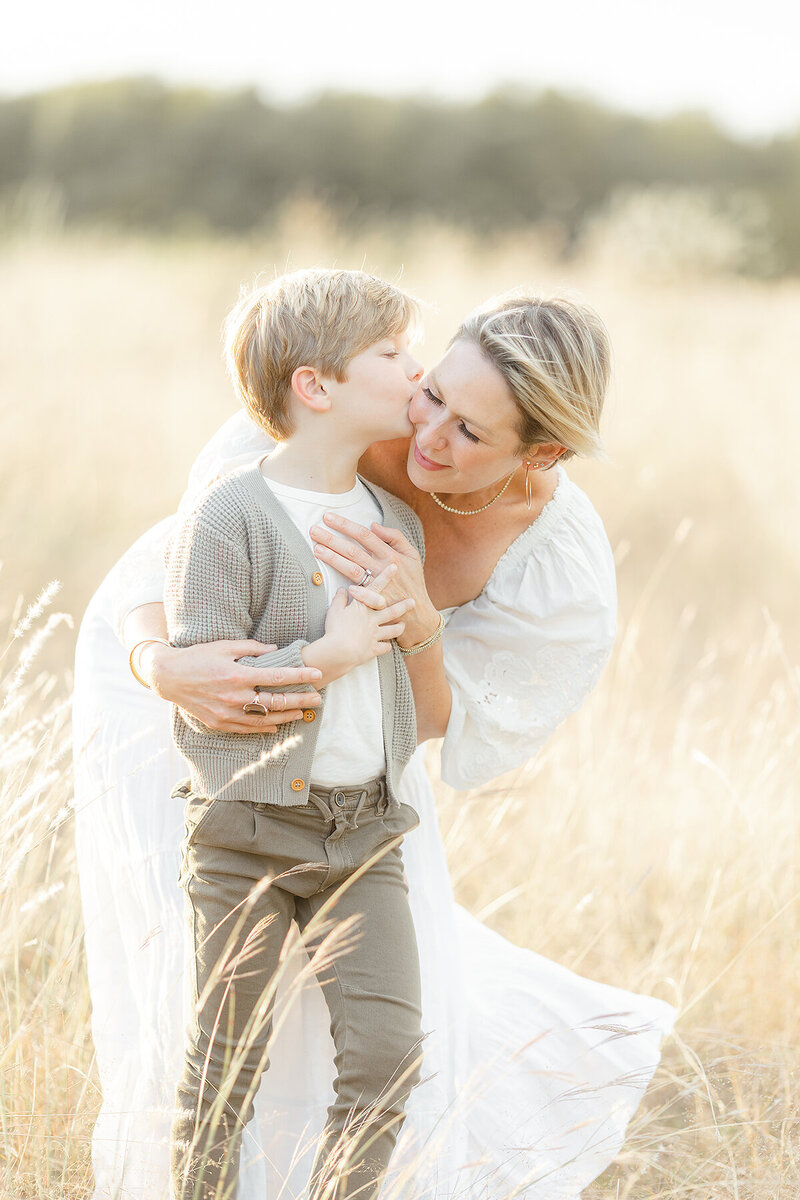 A photo of a mother and her young son standing in a Fort Worth TX park in the middle of a field. She is holding him close as he kisses her cheek.