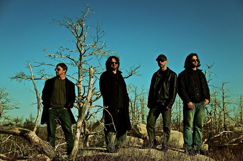 Band portrait Tri City Villains four members wearing coats and sunglasses standing amid sticky trees blue sky behind them