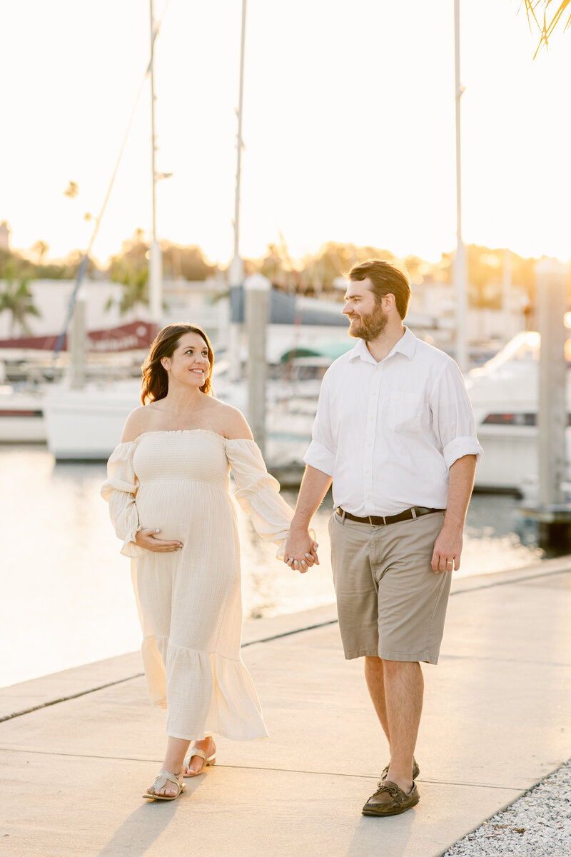 Couple walking along Tampa marina at sunset while holding hands and pregnant mother holds baby bump in stunning white dress