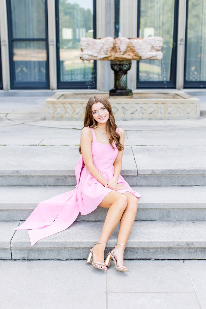 senior girl posing on steps with pink boutique dress on, gold heels and dark brown long hair
