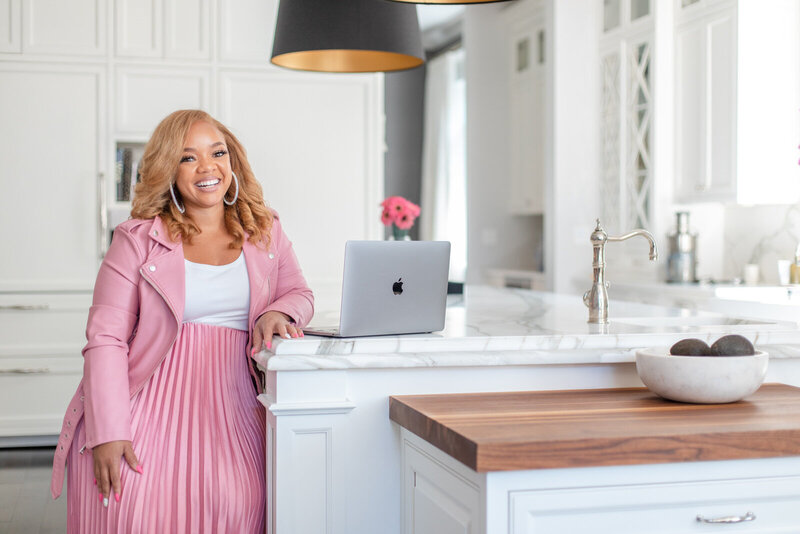 african american woman in pink jacket leans arm on kitchen counter next to a grey macbook pro laptop