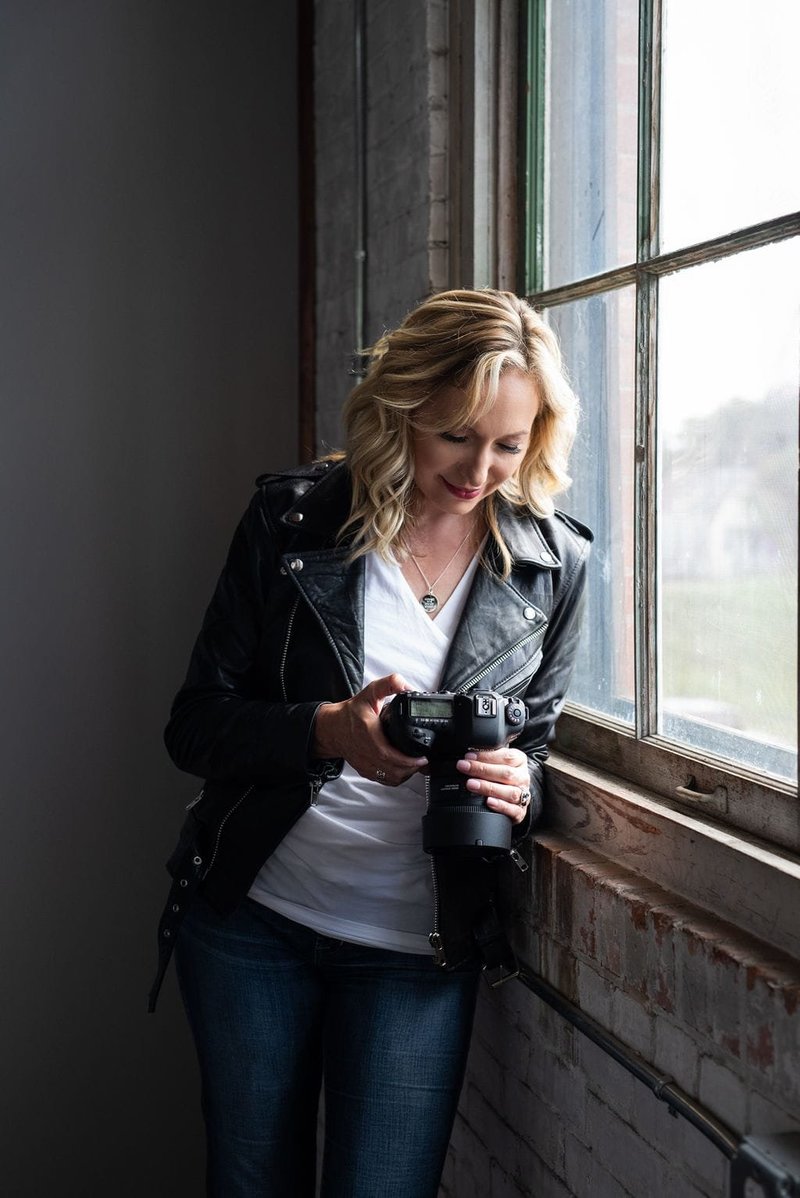 Jen Denton holding a camera looking at the back of it standing next to a window