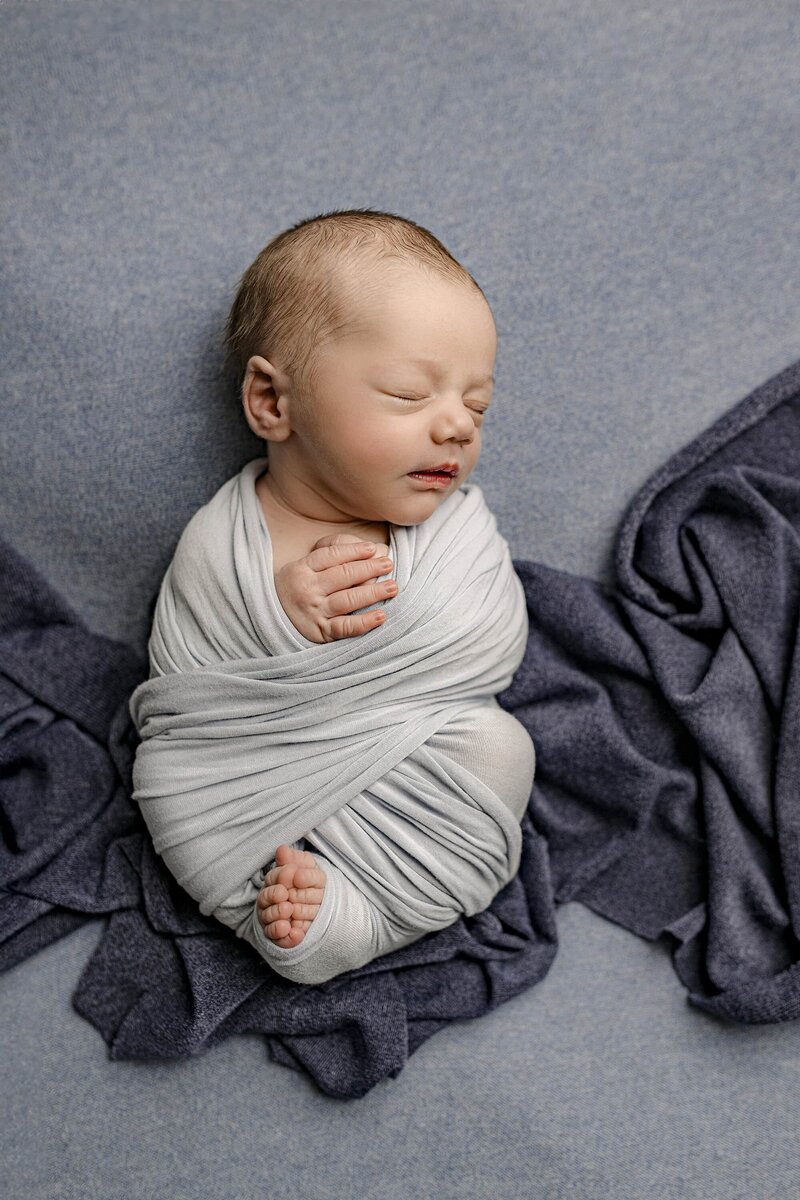 Charlotte Area newborn boy is wrapped and a blue wrap with his face turned to the side laying on a blue background