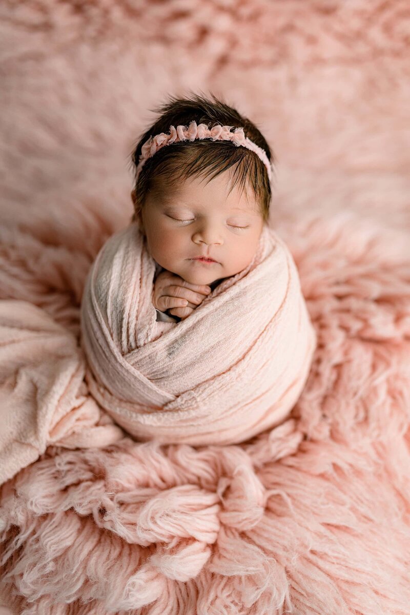 Dark hair is the feature of this image as a newborn baby girl is wrapped in pink on a pink layer wearing a tiny headband and her hands folded gently under her chin