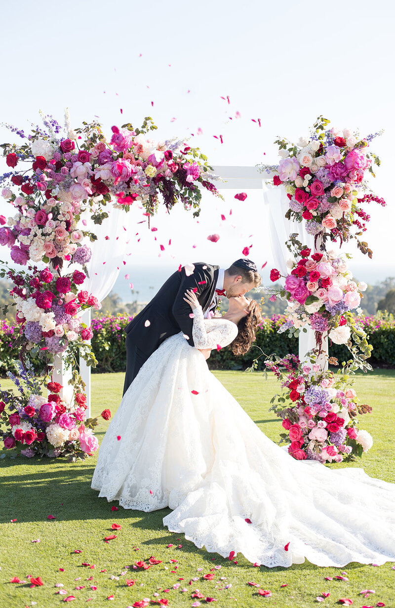 This wedding arbor holds bouquets of red, purple and shades of pink flowers with the contrast of the bride and groom in front. This wedding was held at the Montecito Country Club in Santa Barbara, California.
