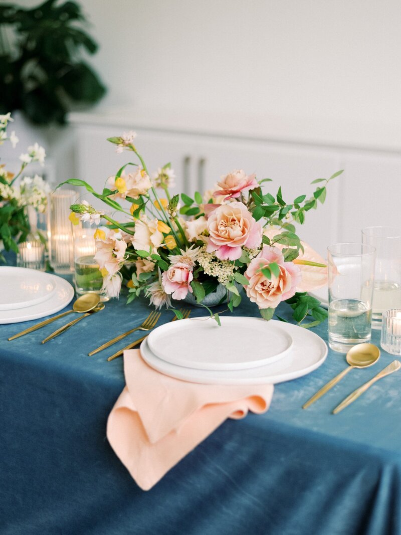 An Anthousai floral arrangement with pink and yellow flowers on a table with a blue velvet tablecloth.