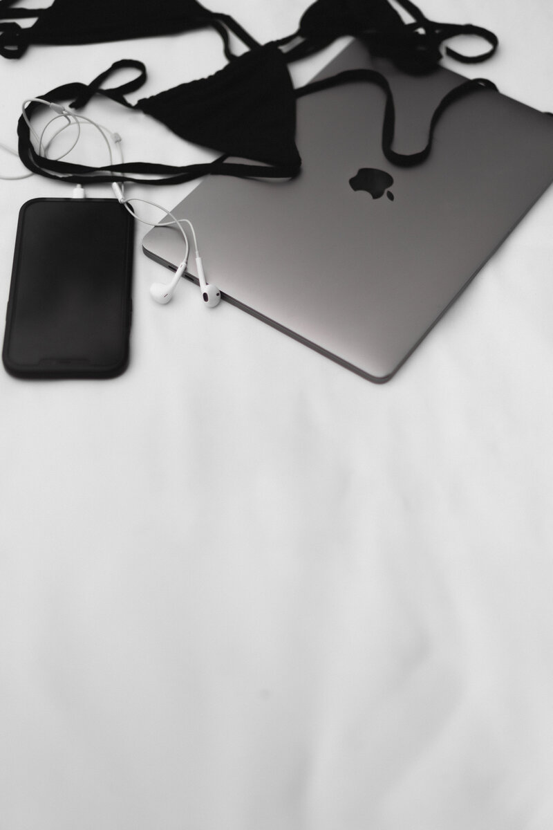 Apple laptop sitting on a bed with a phone and black bikini