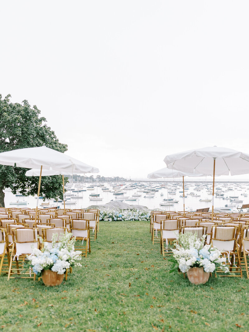 Outdoor empty wedding ceremony space with chairs, umbrellas, and floral arrangements in front of the water and boats