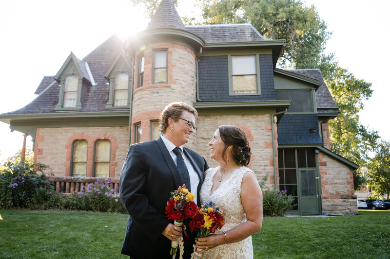 LGBTQ brides smiling at each other while holding bouquets in front of a historic stone mansion during their Fort Collins wedding.