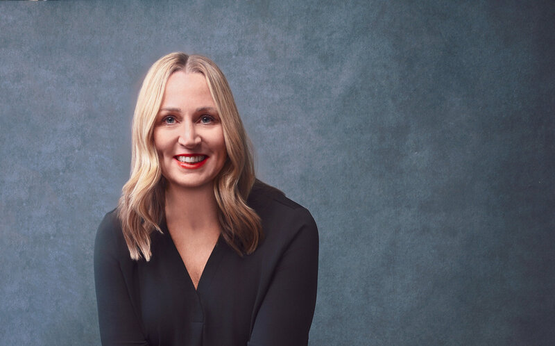 Jenn Cloutier keynote speaker headshot in black blouse with confident smile and professional pose against textured blue background.