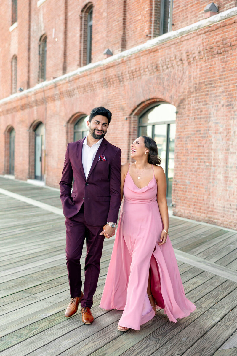 A couple walks hand in hand along a wooden boardwalk. The man wears a burgundy suit, and the woman wears a flowing pink dress. They are smiling and appear joyful, set against the backdrop of a brick building.