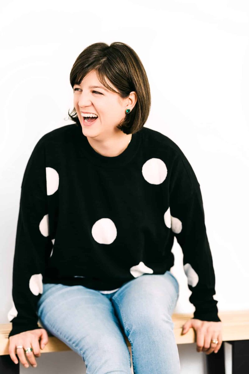 Woman in polkadot sweater sitting on bench with white background.
