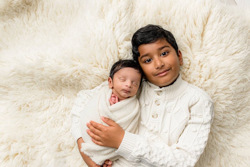 Older brother is dressed in a cream sweater with his newborn baby brother wrapped in cream laying beside him on a fur rug both of their eyes are closed