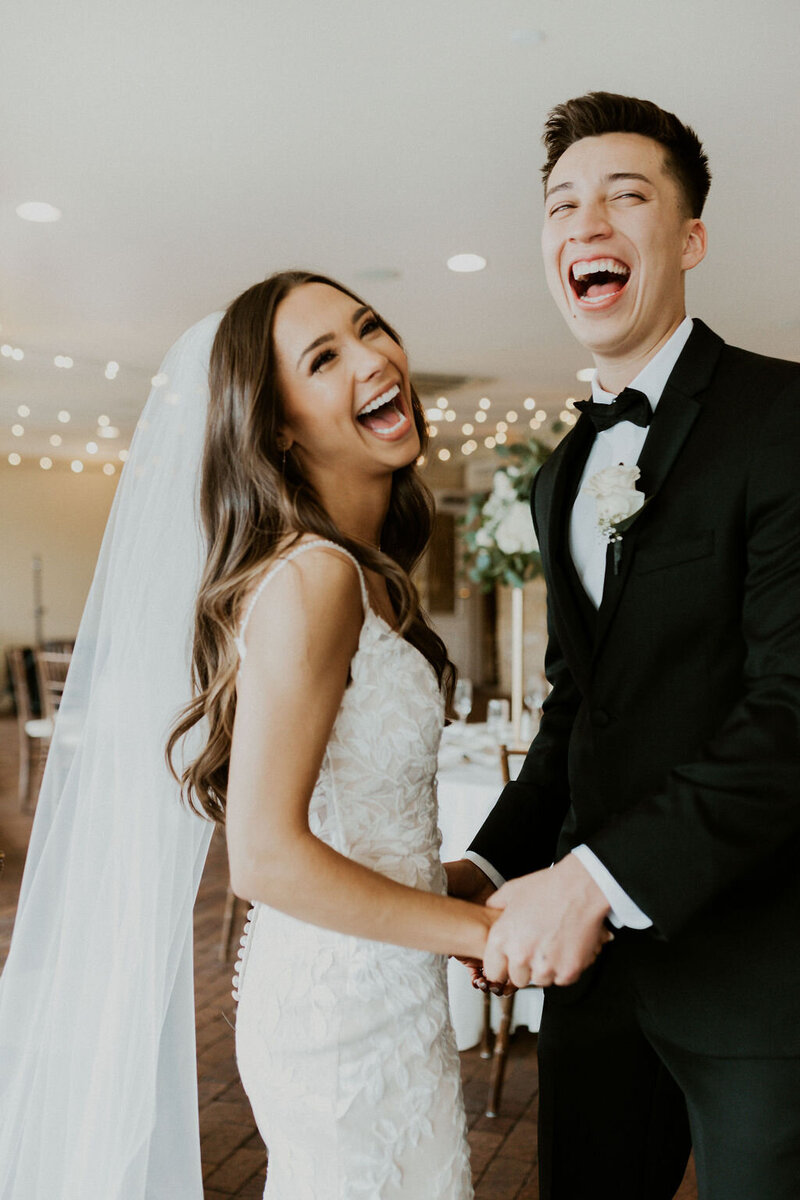 Bride and groom facing each other, holding hands and laughing towards camera