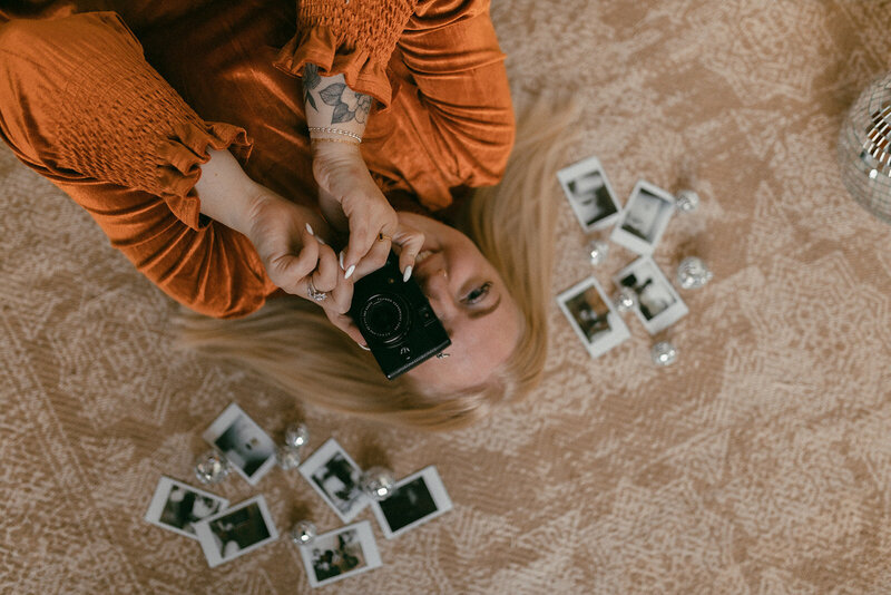 Vintage, moody lifestyle branding headshot of Morgan Ashley Lynn Photography Owner Morgan. Morgan is laying on a rug looking up at the camera with her own camera taking a picture with polaroids around her on the ground wearing an orange shirt.