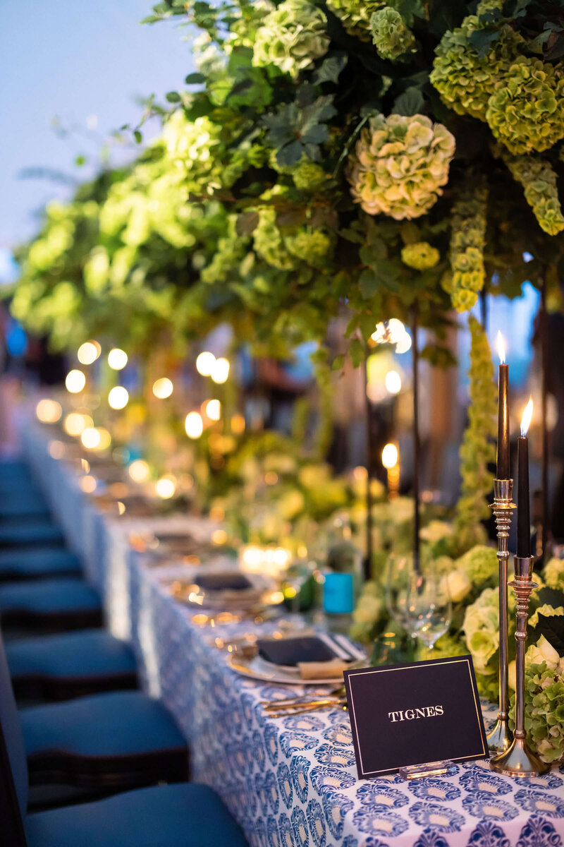 luxury candlelit event dinner on long tables with blue chairs blue patterned linen and tall green flowers