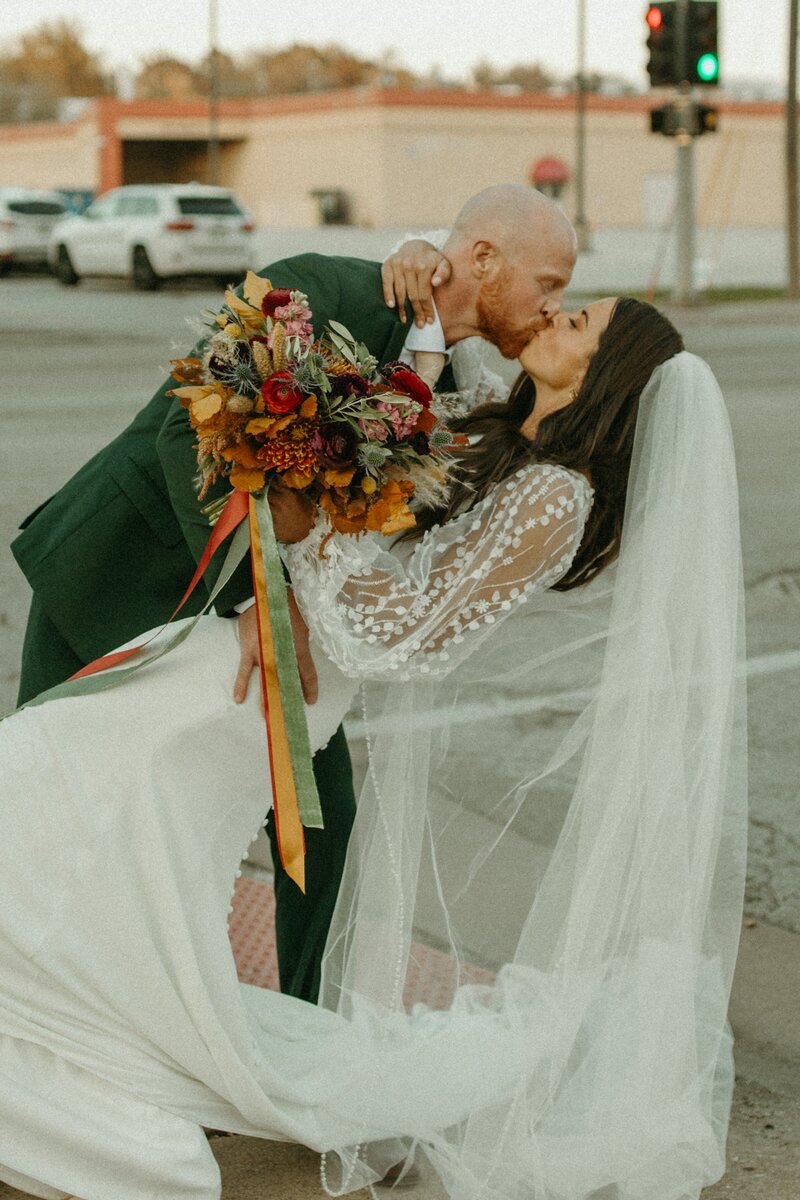 A bride and groom kiss on a city street in Davenport; the bride holds a large bouquet while her veil flows down.