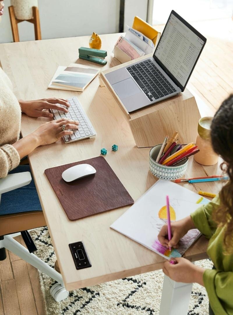 Mom entrepreneur working on laptop while child colors at the desk.