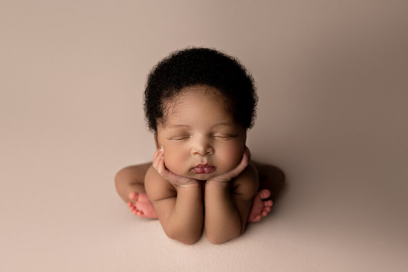 Katie Marshall, best newborn photographer in New Jersey, captures a black newborn baby in froggy pose. Her hands are under his cheeks and has a full head of curly hair. The artful lighting shows lots of depth and shadows. The baby is bare against the blush backdrop.