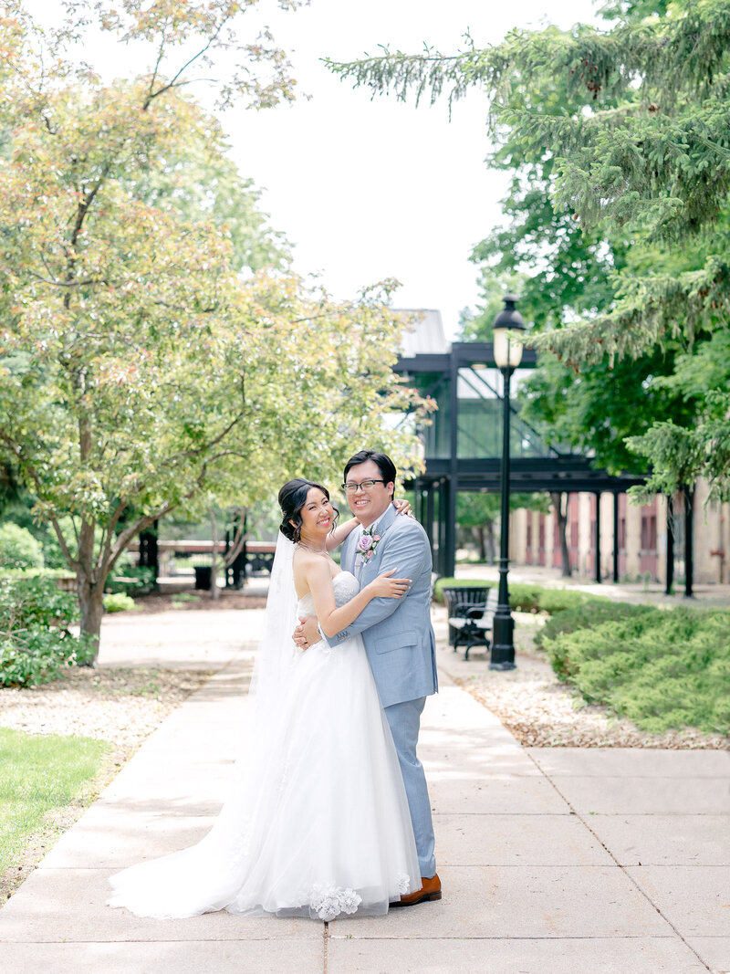 Bride and groom smiling with their arms around each other standing on a sidewalk with trees around them