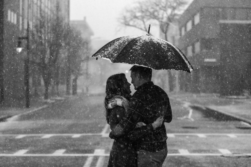 Engaged couple cuddling under an umbrella as snow falls around them on Gannon University's campus in downtown Erie, PA