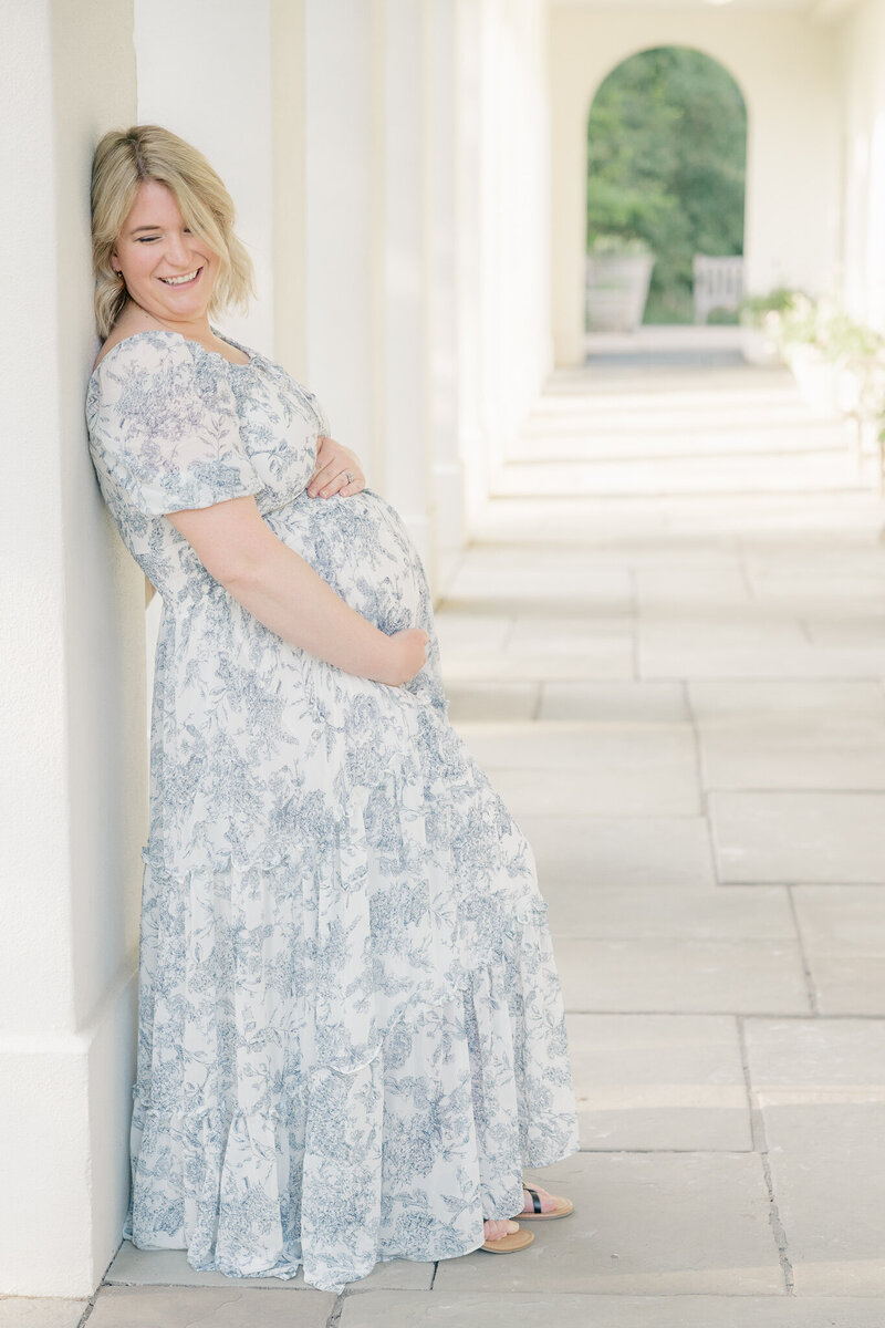 Pregnant mother in a blue floral dress leans against a pillar at Newfields while holding her baby bump and smiling, Indianapolis Photographer