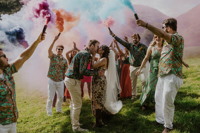 Bride and groom kissing while surrounded my the rest of the bridal party who are holding up coloured smoke grenades