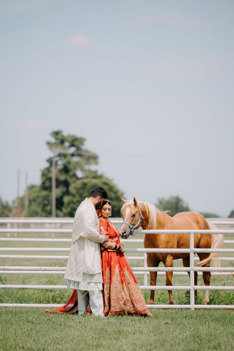 A couple, dressed in traditional Indian wedding attire (the woman in a red and gold lehenga, the man in a cream-colored kurta), stands near a white fence in a grassy field. A light brown horse is also visible in the image, standing next to the couple.  The background features greenery and a partly cloudy sky.
