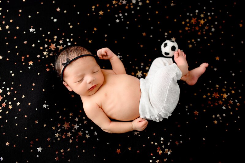 newborn in grey pants is posed on a black background with a soccer baall
