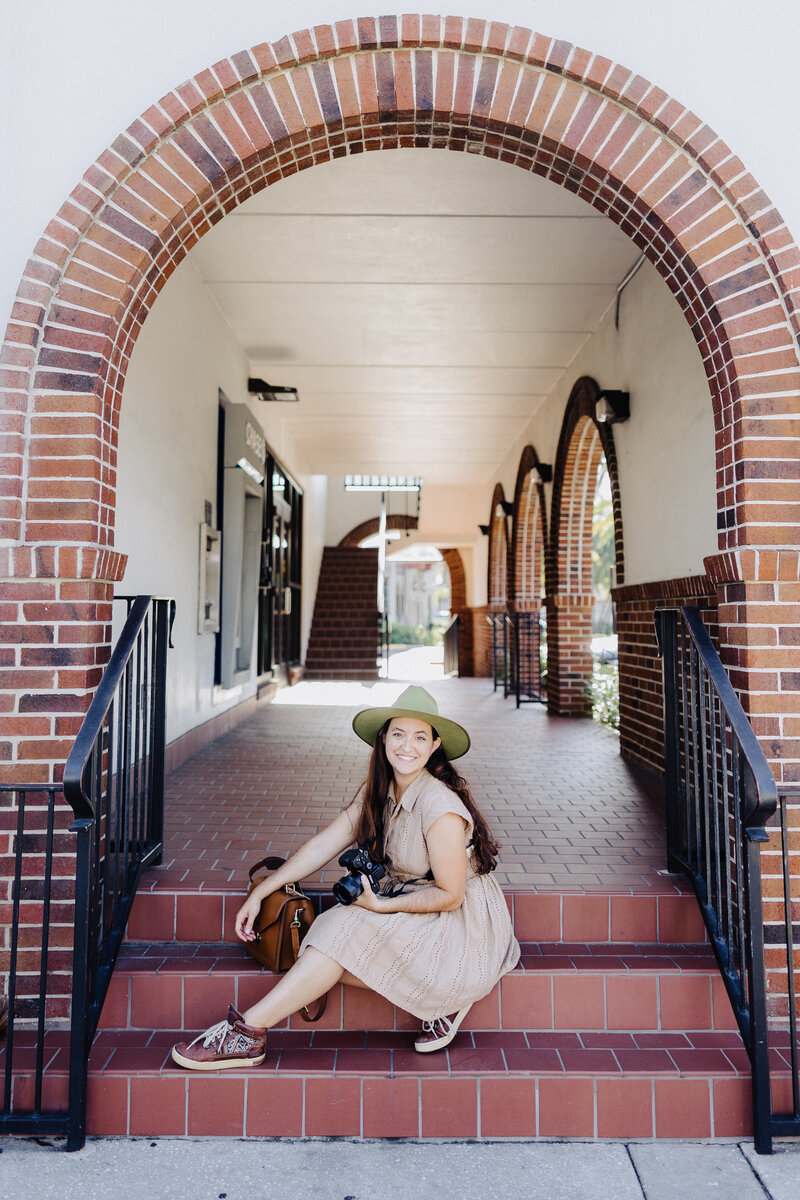 Orlando Wedding Photographer Jo of Four Loves Photo and Film is posed with her camera, sitting on the steps of a beautiful brick and stucco building with arched doorways.
