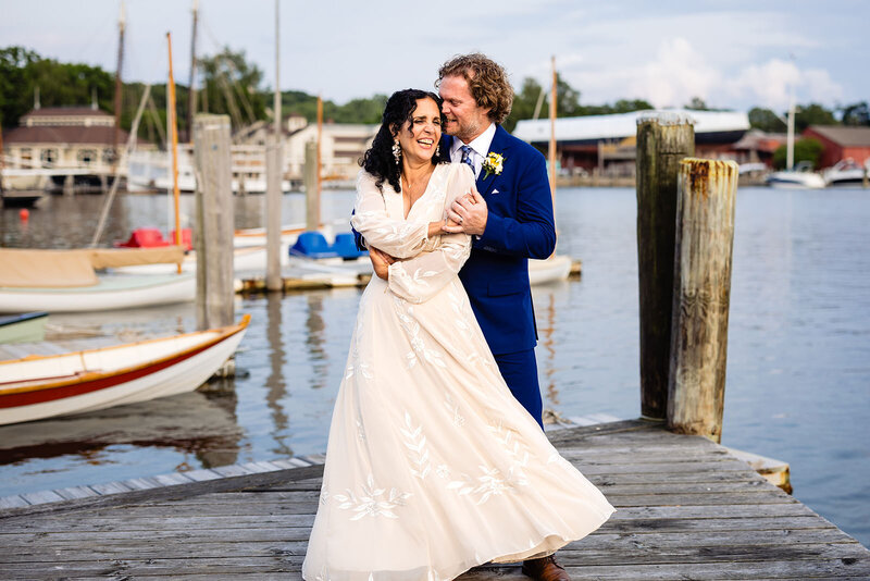Bride and groom embracing on a wooden dock at Mystic Seaport, with sailboats and calm waters in the background