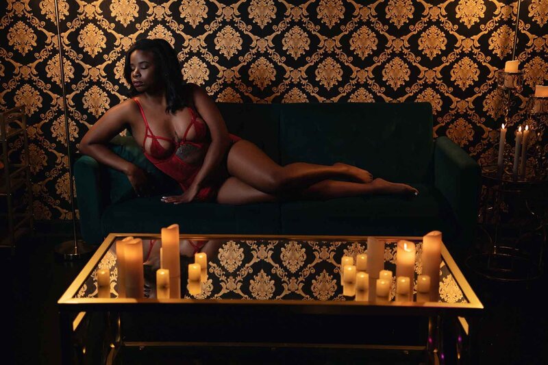 Dark skinned woman posing on emerald velvet couch infront of black and gold baroque backfrop