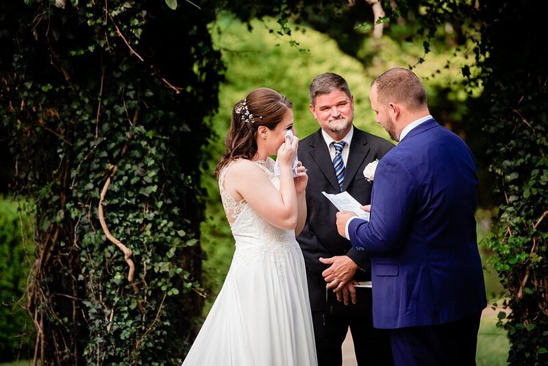 Bride using tissue to dab at tears during ceremony near an ivy wall