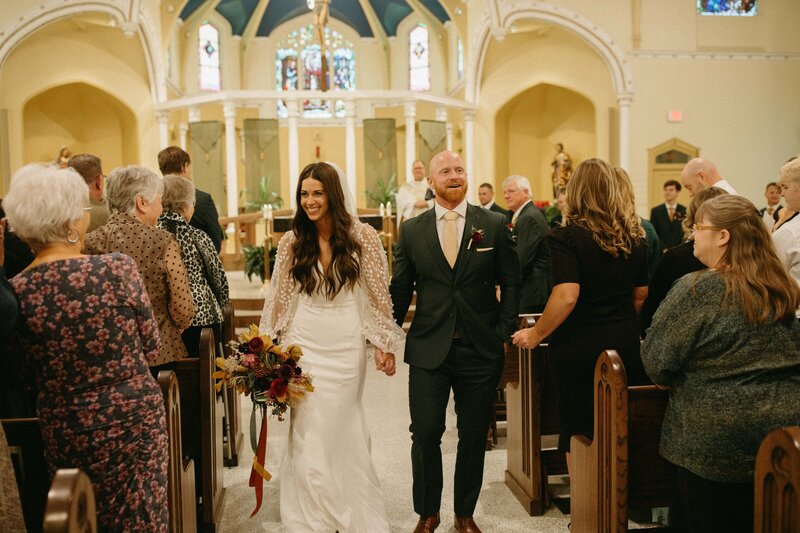 A bride and groom joyfully walk down the aisle of a church in Iowa after their wedding ceremony, surrounded by guests.