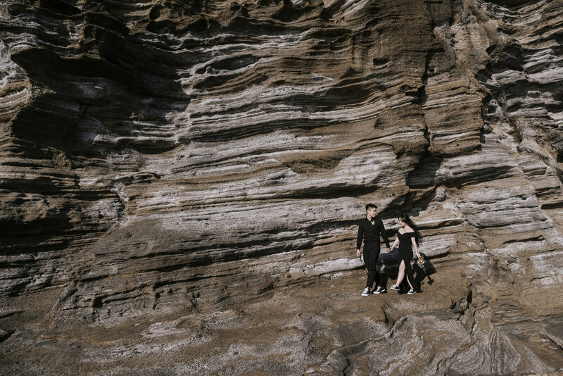 couple dressed in black walk along the rock formations at Yongmori Coast during their pre-wedding