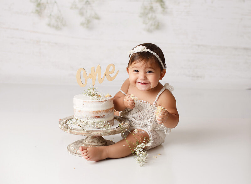 one year old baby girl celebrating her first birthday a her cake smash session wearing a vintage lace romper and matching hair bow