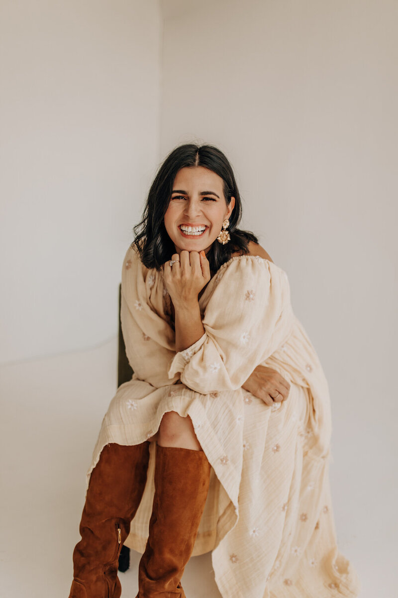 Bianca Flanagan, a San Antonio photographer, sitting in front a cream-colored backdrop at a studio. She is smiling in a cream dress while taking branding images.