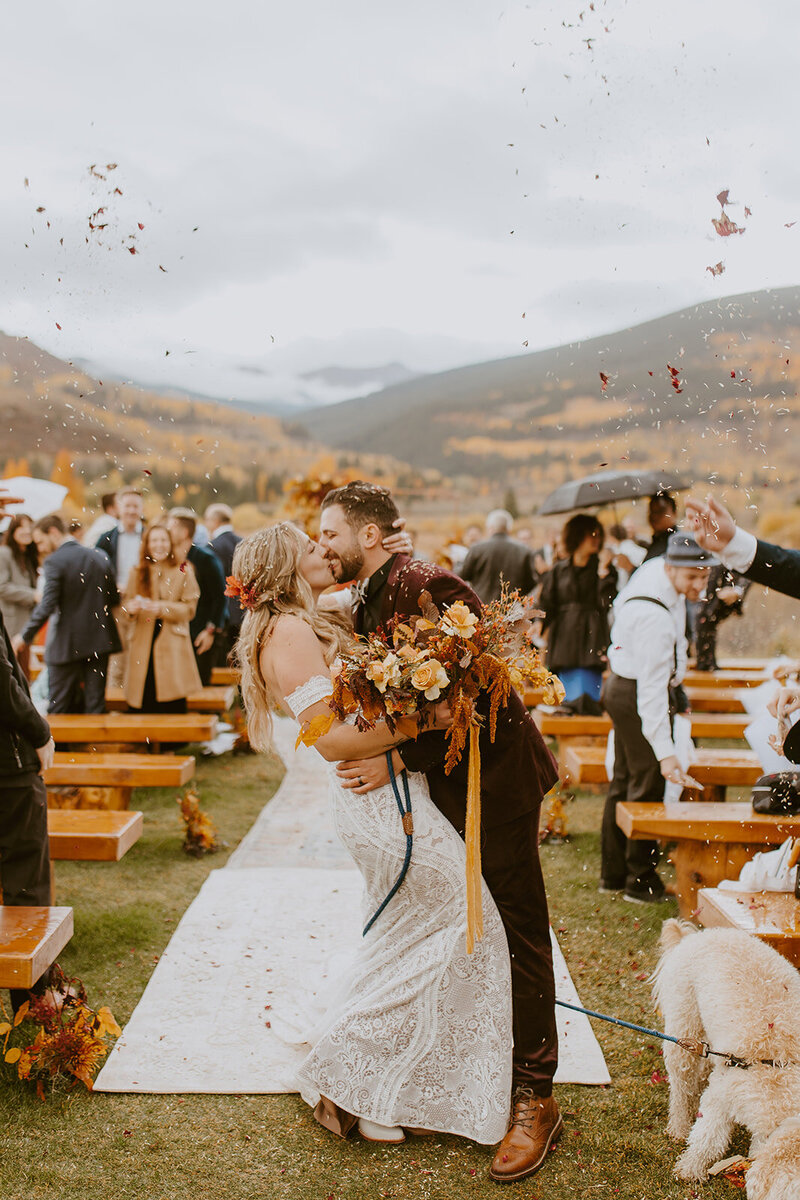 Bride and groom share a kiss in front of a mountain backdrop while  guests throw confetti.