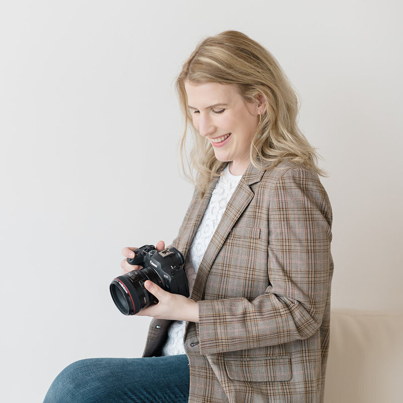 Photo of Shaunae Teske, a newborn photographer in Seattle, wearing a plaid blazer looking down at her camera and smiling while sitting on a stool