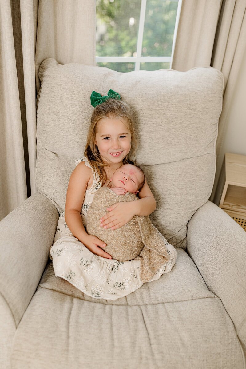 toddler sister dressed in green flowers holds her newborn brother wrapped in brown as they sit in a chair