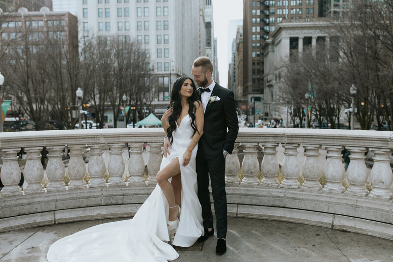 Bride with long, brown, curly hair leans against tall groom in a Chicago park in front of barren trees and skyscrapers.