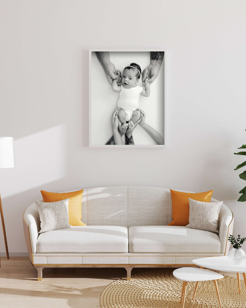 A modern living room with a white sofa featuring yellow and beige pillows, a round wicker coffee table, and a large framed black-and-white photo by a Jacksonville newborn photographer hanging above the sofa.