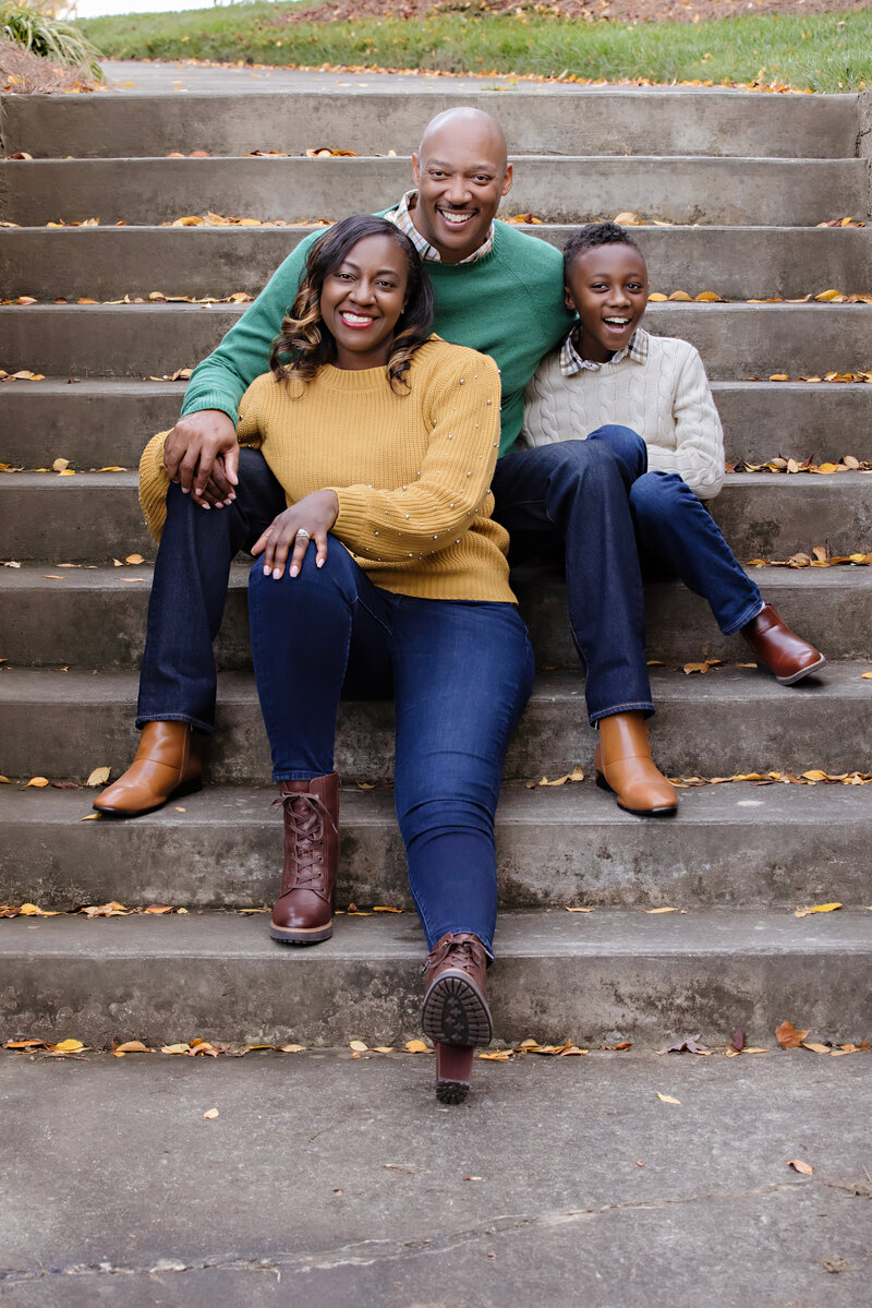 Ballantyne's Best family photographer captured this portrait of a family on a concrete staircase in lovely coordinated fall inspired outfits.  Dad wears a green sweater over a plaid button down shirt, mom has a beautiful golden sweater with jeans and their son is wearing an oatmeal sweater over a plaid shirt. The button down shirts help tie the sweater colors together. 