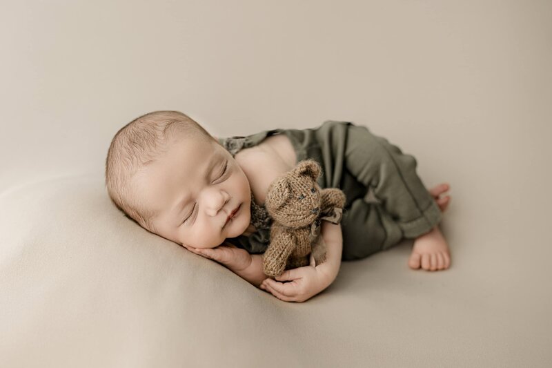 Charlotte Area infant cradles a stuffed bear while laying on his side with his other hand underneath his cheek dress and dressed in green overalls