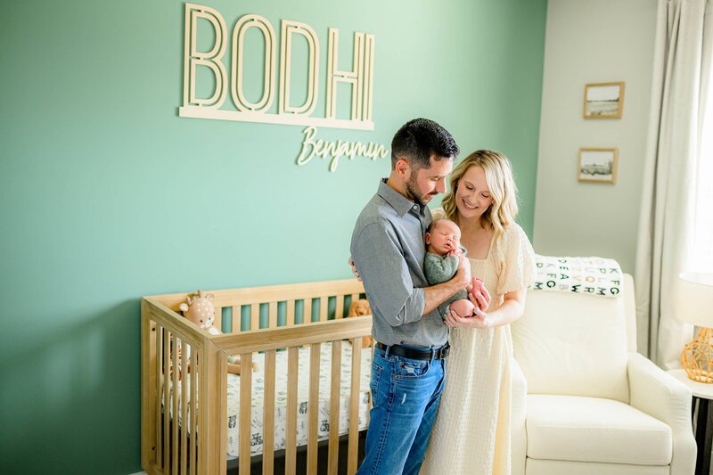 father and mother hold newborn son in his nursery with his crib behind them