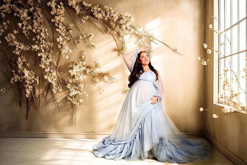 Pregnant Charlotte area mother stands in a window studio set up wearing a blue dress with one of her hands above her head