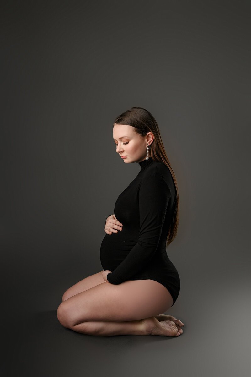 dark hair pregnant mother sits on ground wearing a black bodysuit on a grey studio background cradling her baby bump