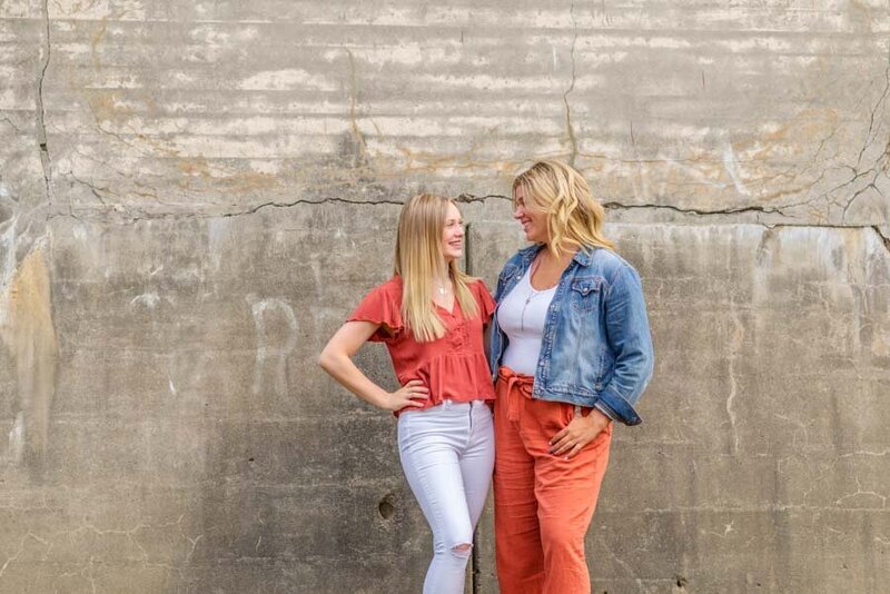 Two women smiling and talking, standing in front of a textured concrete wall, one in a red blouse and white pants, the other in an orange jumpsuit and denim jacket.