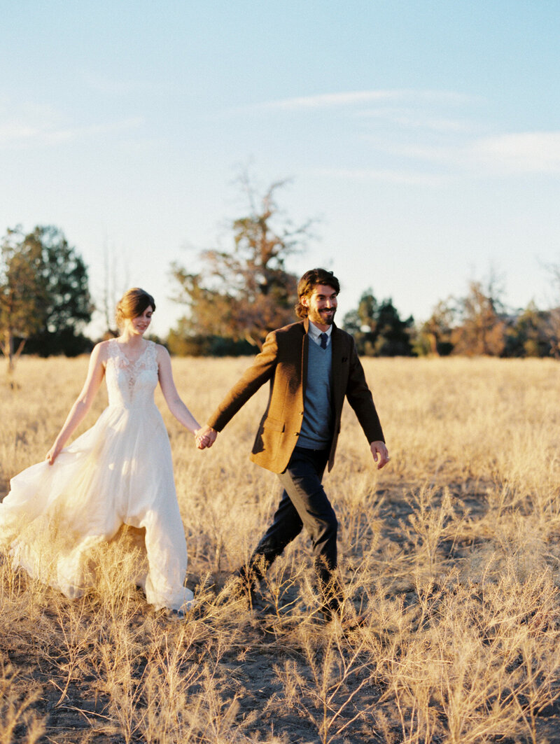 Groom holds his bride's hand and guides her through dried brown grassland in wedding attires.