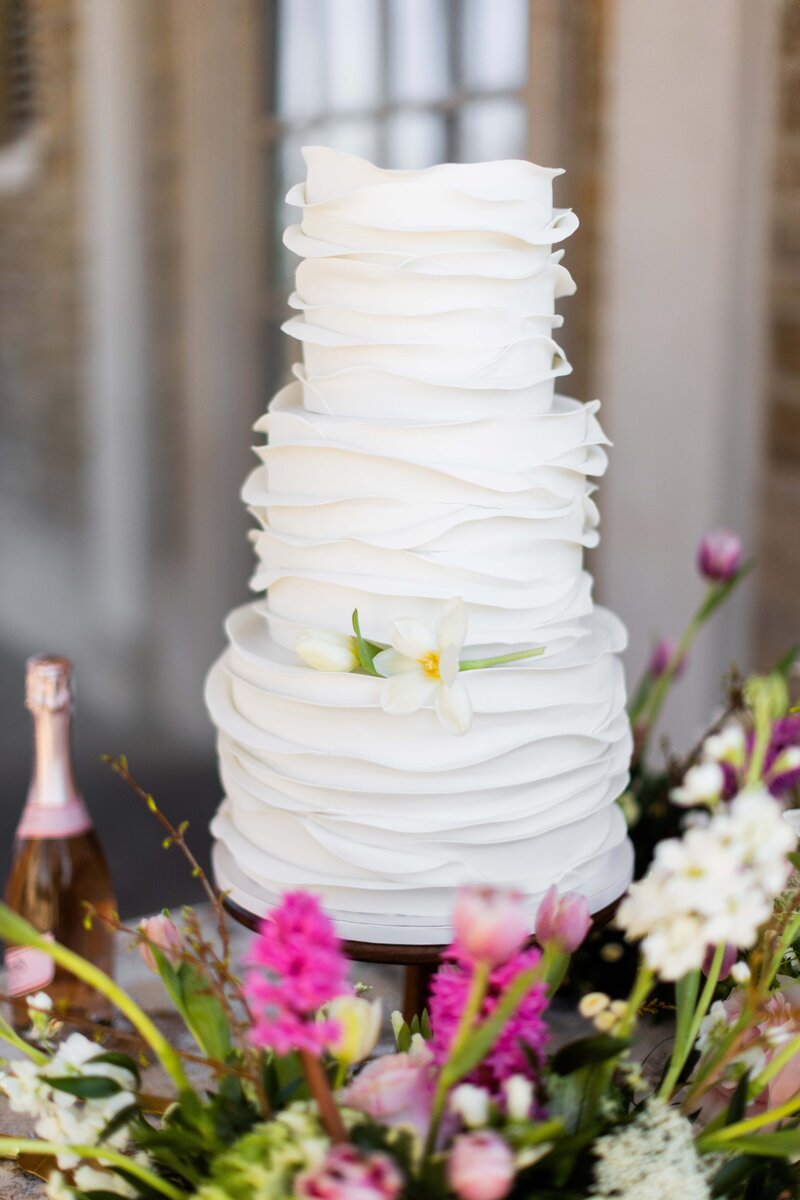 Behold the artistry and elegance of this breathtaking wedding cake crafted by Spoon Fulla Suga, featuring delicate white fondant ruffles that cascade gracefully around each tier. This masterpiece combines exquisite craftsmanship with timeless style, making it a perfect focal point for any wedding reception. The intricate ruffles add a touch of sophistication and luxury, ideal for couples who desire a wedding cake that is both visually stunning and tastefully refined.